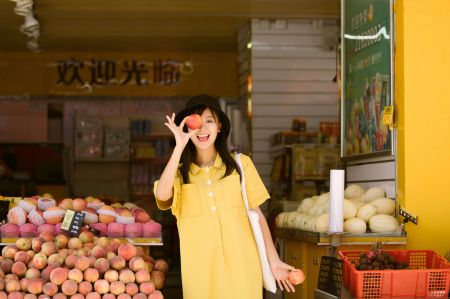 女孩在水果摊前手持桃子，背景为欢迎光临的店铺