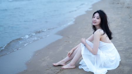 穿着白色连衣裙的美女坐在沙滩上，平静的海面