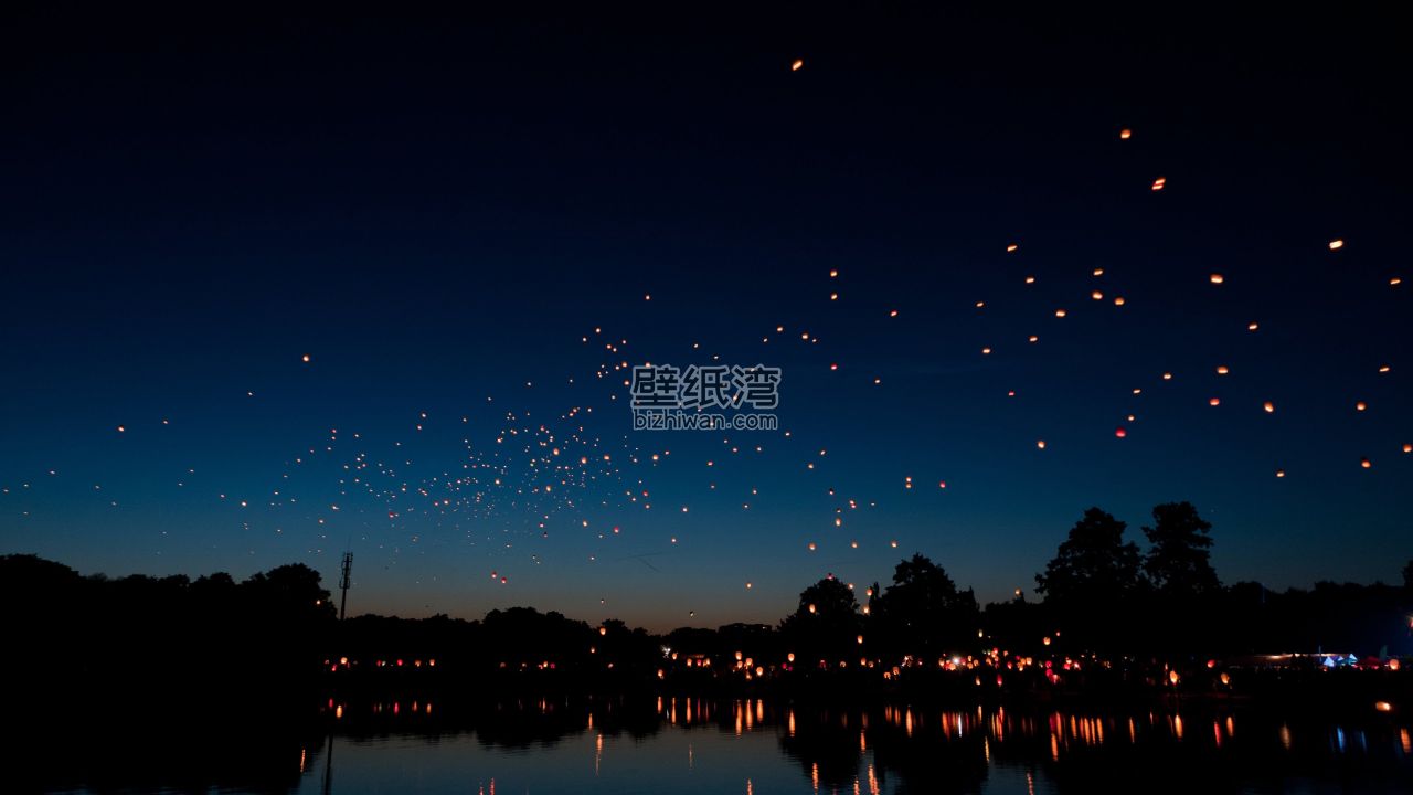 夜晚的天空中飘浮着许多孔明灯，湖面倒映着灯光和树木的轮廓